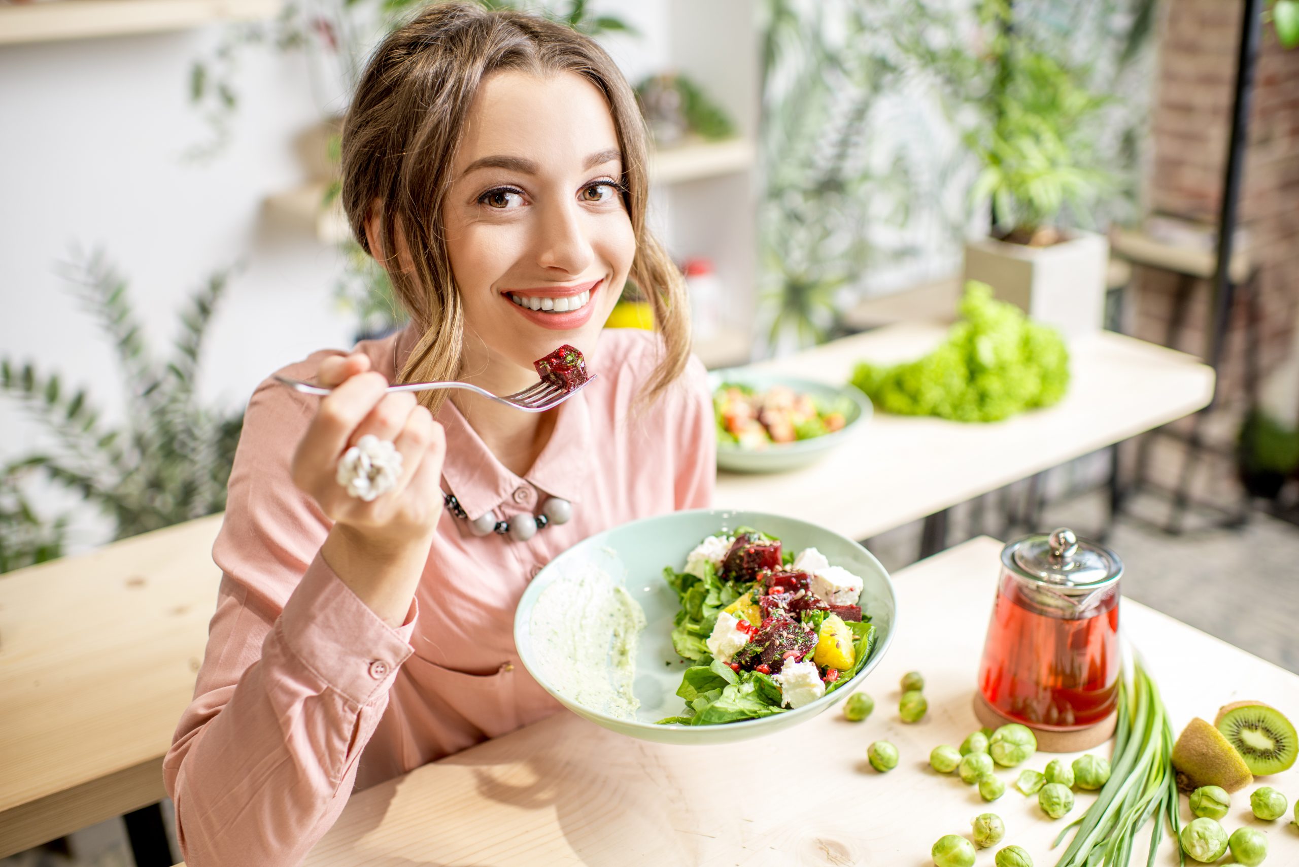 Young woman eating healthy food sitting in the beautiful interior with green flowers on the background