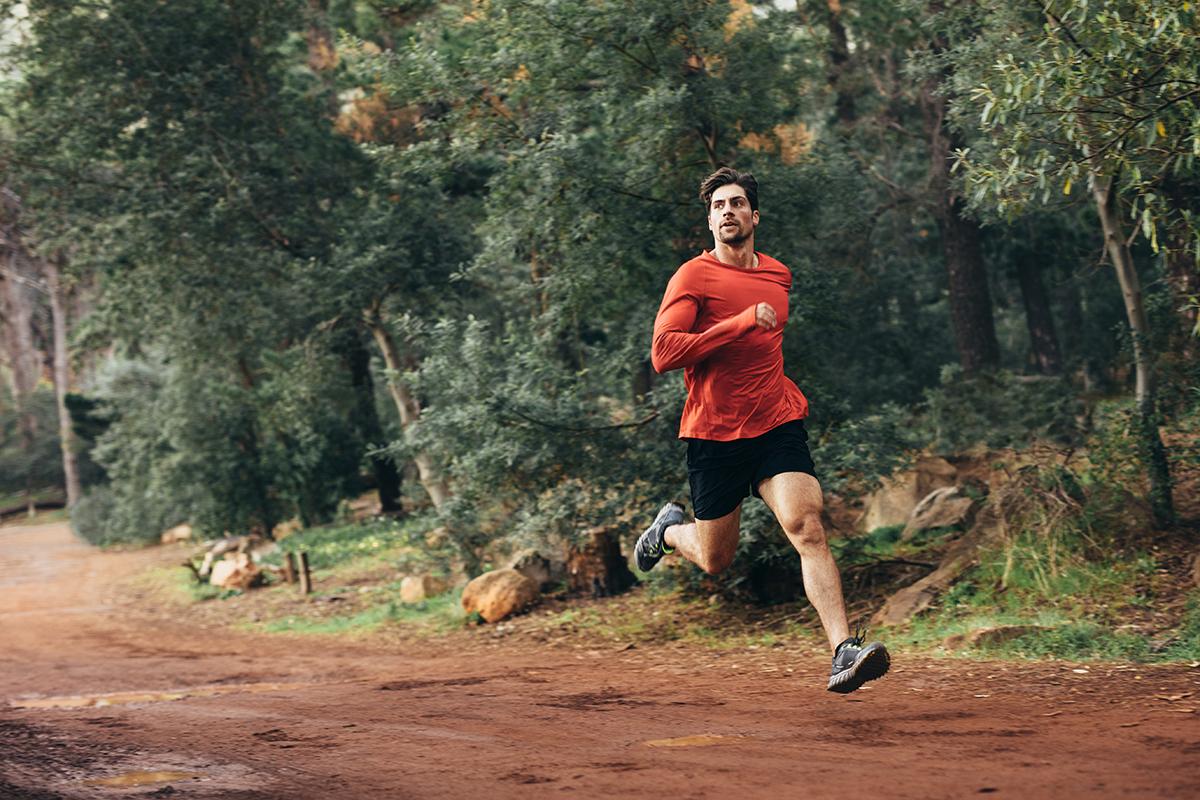 Man running on mud track. Athlete running fast in a park with dense trees in the background.